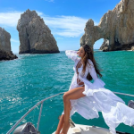 Wanita memakai beachwear hijau stylish di tepi laut dengan latar batu karang dan langit biru cerah